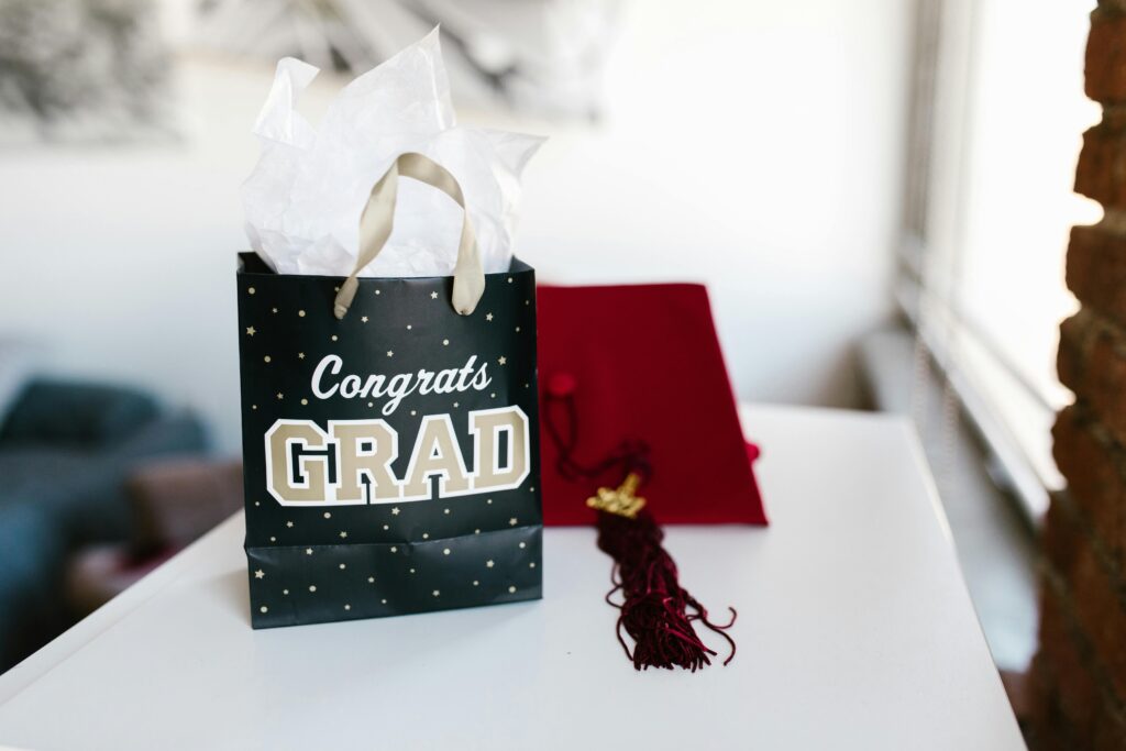 A stylish graduation gift bag alongside a red mortarboard hat on a white table.